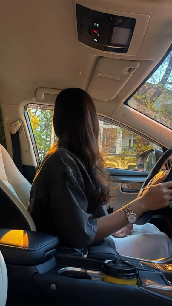 Woman driving a car, looking out the window at a sunny street, with a yellow coffee cup in the console, wearing a watch and denim shirt.