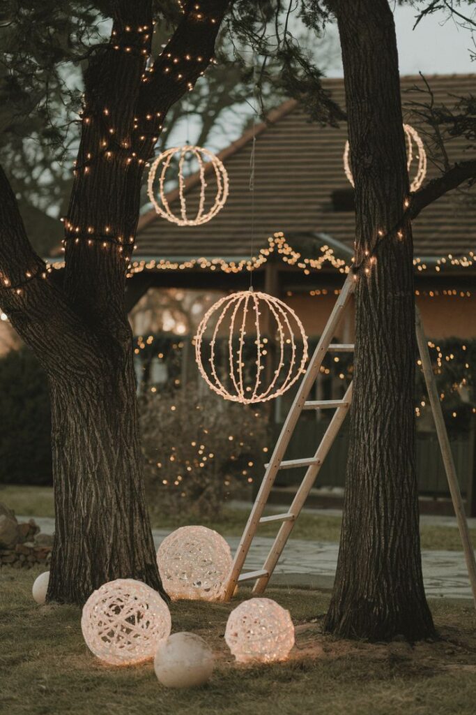 0ecce00c3f330fa315844fae53d429b1 Festive outdoor holiday decorations with glowing orbs and lights in trees, ladder leaning on trunk, cozy garden ambiance at dusk. | Sky Rye Design Festive outdoor holiday decorations with glowing orbs and lights in trees, ladder leaning on trunk, cozy garden ambiance at dusk.
