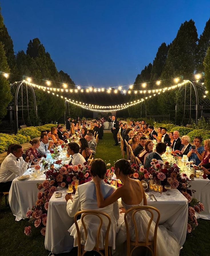 _ 1 Outdoor wedding reception at dusk with string lights, elegantly decorated tables, and guests enjoying the celebration in a garden setting. | Sky Rye Design Outdoor wedding reception at dusk with string lights, elegantly decorated tables, and guests enjoying the celebration in a garden setting.