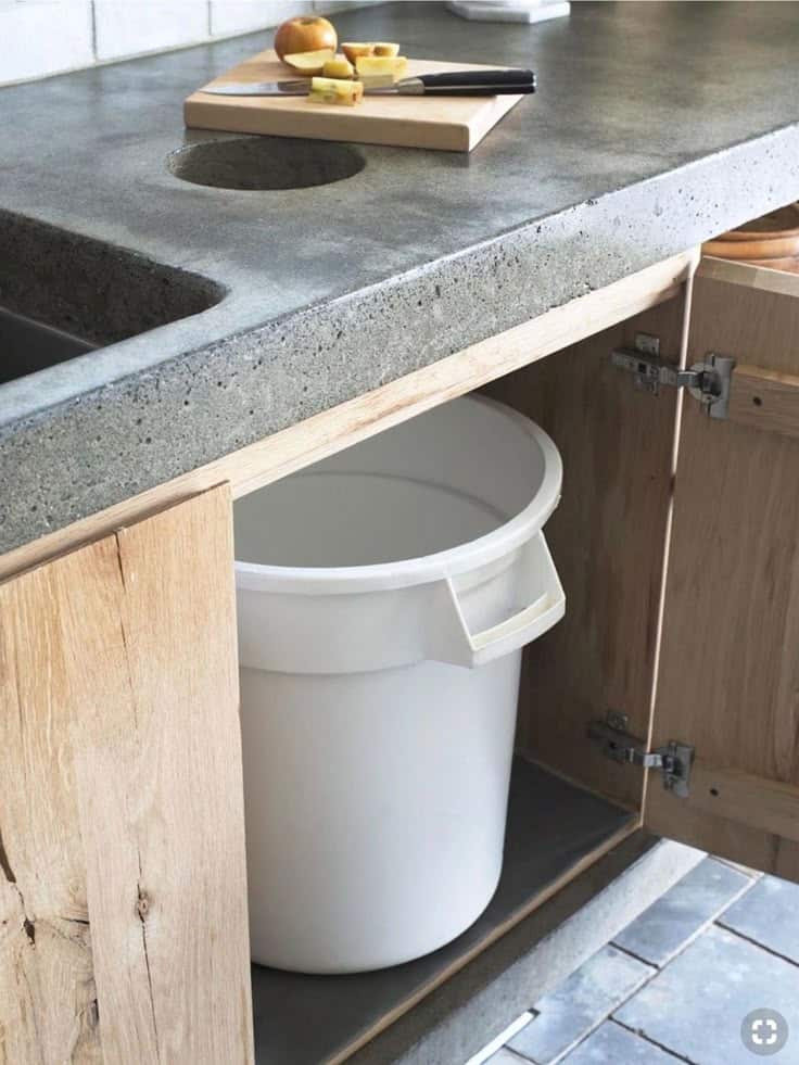 Modern kitchen with a concrete countertop, cutout for waste scraps, and wooden cabinets. White trash bin below for efficient waste disposal.