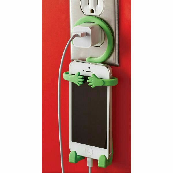 _ 1 Green smartphone holder with arms and legs secures a phone while charging on a wall socket against a red background. | Sky Rye Design Green smartphone holder with arms and legs secures a phone while charging on a wall socket against a red background.