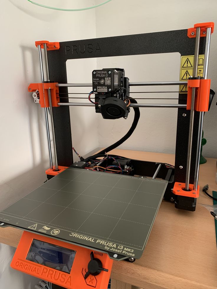 3D printer setup on a desk; Original Prusa i3 MK3, featuring an extruder and control screen for precise 3D printing tasks.