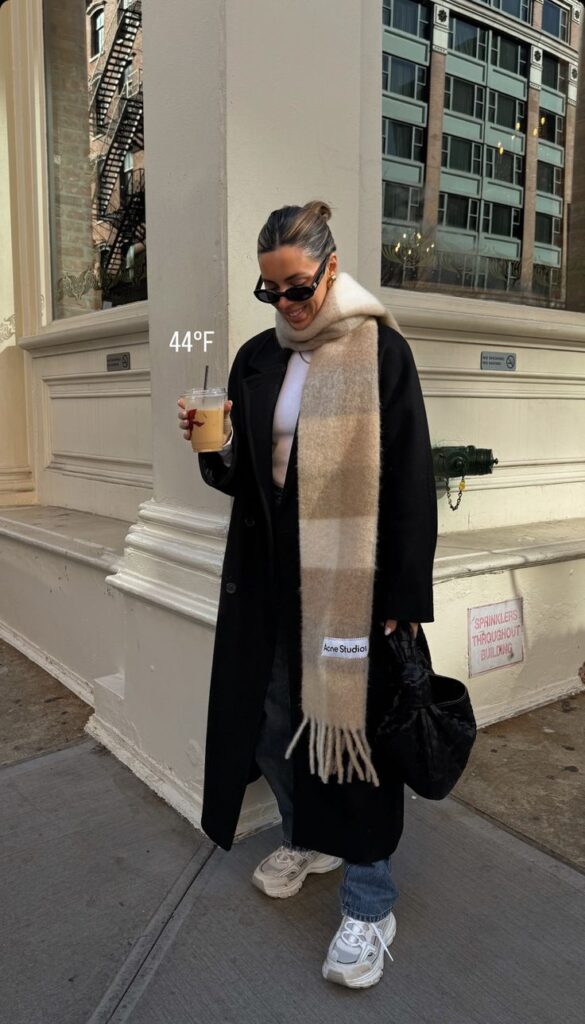 Woman in winter outfit holding iced coffee, wearing a long scarf and sunglasses, walking on a city street, 44°F.