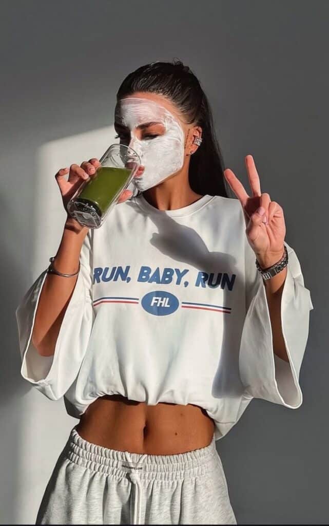 _ 1 Woman with face mask drinking green juice and flashing peace sign in casual outfit. | Sky Rye Design Woman with face mask drinking green juice and flashing peace sign in casual outfit.