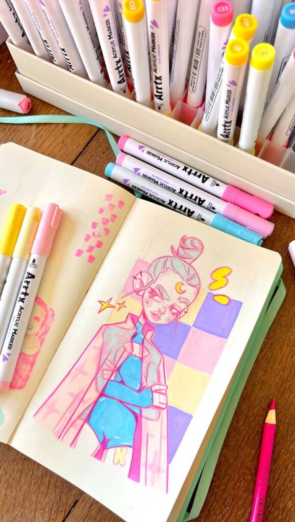 Colorful anime character sketch on notebook surrounded by Artix markers. Creative art supplies on wooden table.