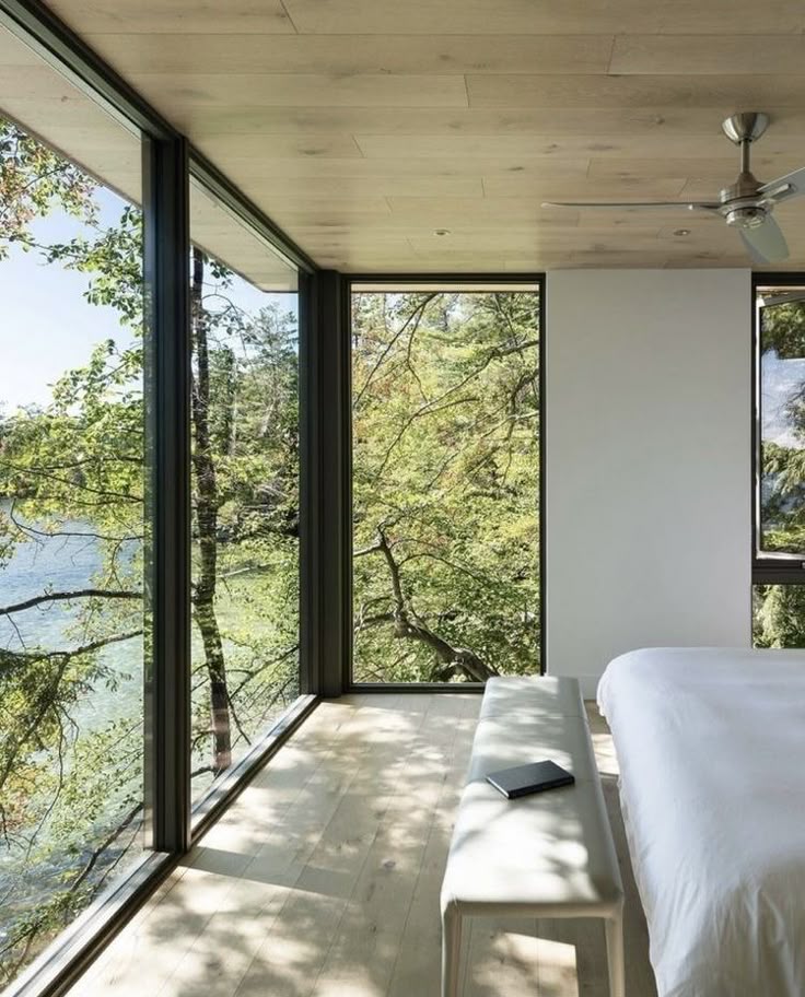 _ 10 Modern bedroom with large windows overlooking a serene wooded lake view, featuring a minimalist bed and ceiling fan. | Sky Rye Design Modern bedroom with large windows overlooking a serene wooded lake view, featuring a minimalist bed and ceiling fan.