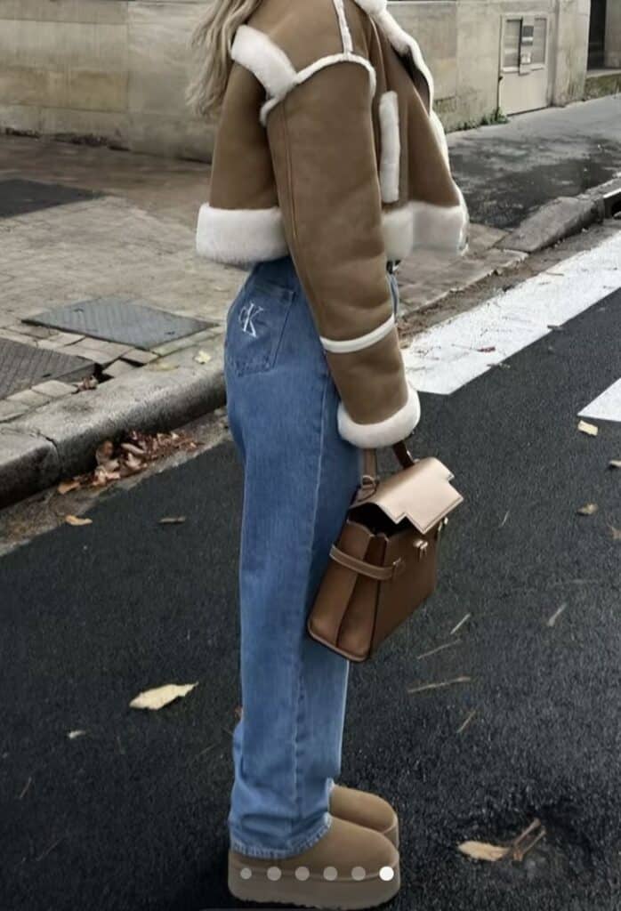 Person in stylish shearling jacket, blue jeans, and brown boots with matching handbag, standing on the street.