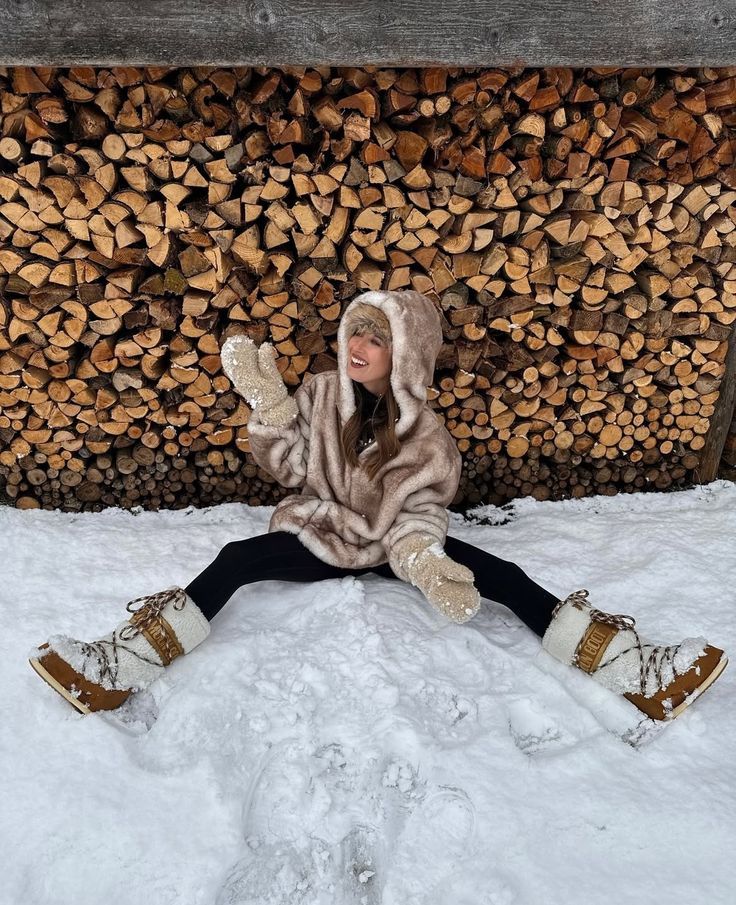 Woman in fur coat and snow boots sits happily in snow, waving with woodpile backdrop. Winter fashion fun.