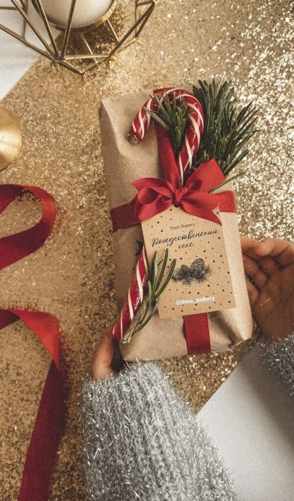 _ 10 Hand holding a festive gift wrapped in brown paper, adorned with a candy cane, red ribbon, and greenery, against a glittery background. | Sky Rye Design Hand holding a festive gift wrapped in brown paper, adorned with a candy cane, red ribbon, and greenery, against a glittery background.
