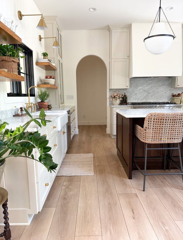 Modern kitchen with white cabinets, wood accents, and greenery, featuring a marble island and wicker bar stool.
