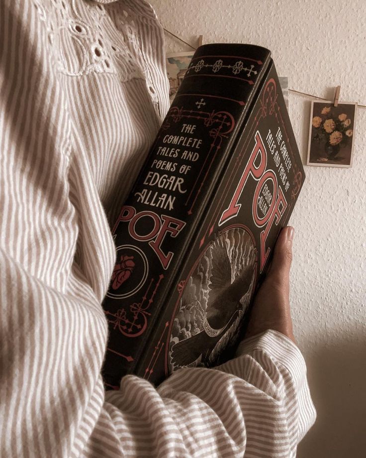 Person holding The Complete Tales and Poems of Edgar Allan Poe book, wearing a striped shirt, with cozy vintage decor in the background.