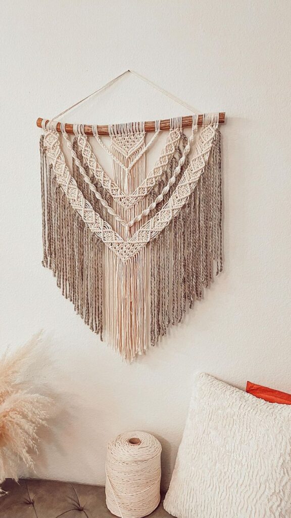 Handmade macramé wall hanging in cream tones, displayed above a cozy couch with textured pillow and yarn spool, creating a boho-chic space.