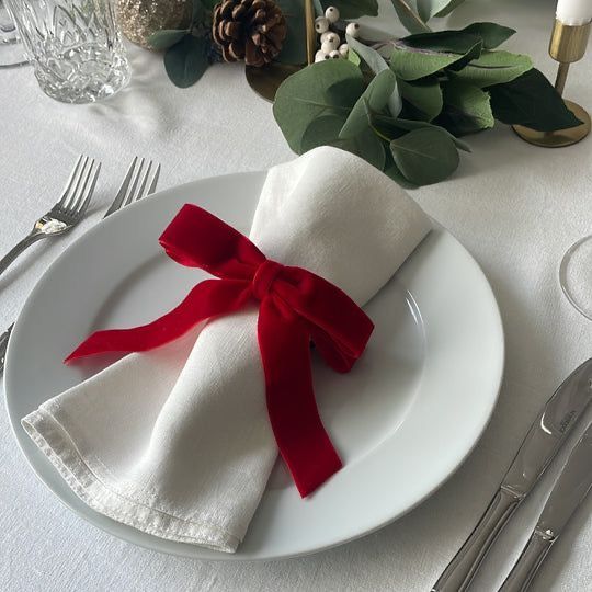 Elegant holiday table setting with white napkin and red bow on a plate, surrounded by greenery and festive decor.