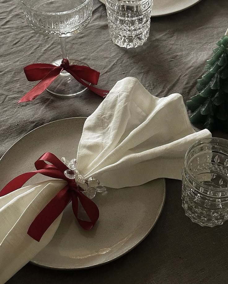 Elegant holiday table setting with crystal glasses, white napkin tied with red ribbon on a plate, and decorative greenery.