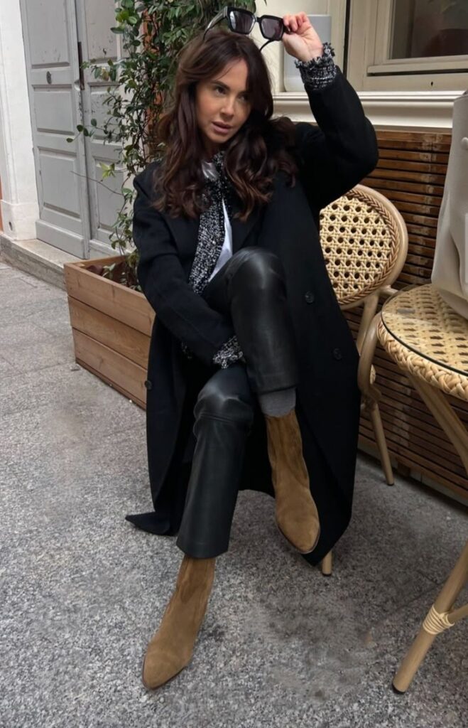 Stylish woman in black coat and leather pants poses outdoors, holding sunglasses above her head near a wicker chair.
