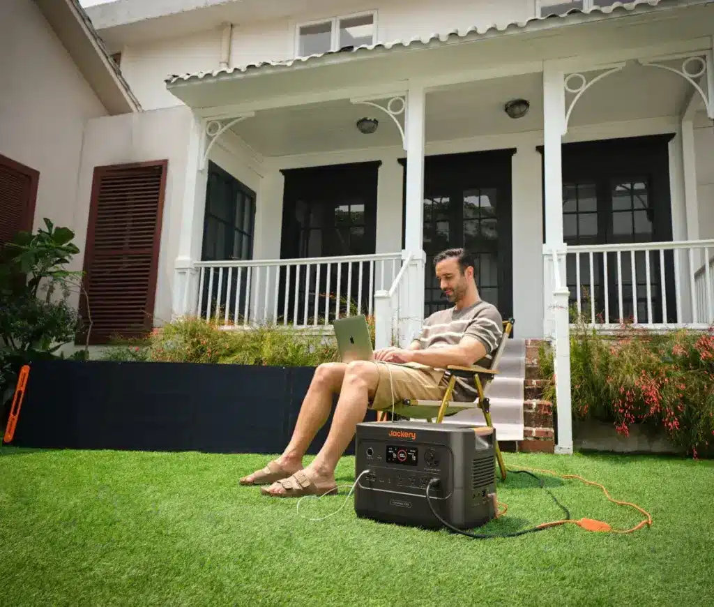 1200_1020_Yard Man working on a laptop in a garden with a portable solar generator in front of a white house. | Sky Rye Design Man working on a laptop in a garden with a portable solar generator in front of a white house.