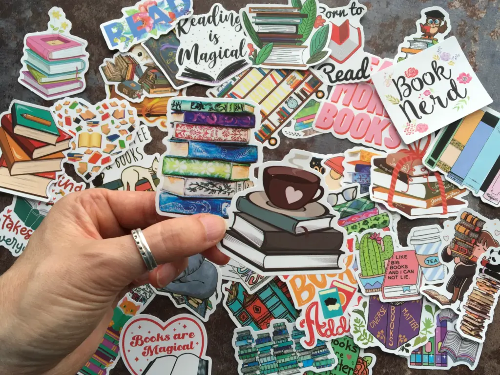 12D82A7A-F103-41DB-8D3E-17385B602AD1_1_201_a Assorted book-themed stickers featuring stacks of books, reading quotes, and colorful designs, held by a hand with a silver ring. | Sky Rye Design Assorted book-themed stickers featuring stacks of books, reading quotes, and colorful designs, held by a hand with a silver ring.