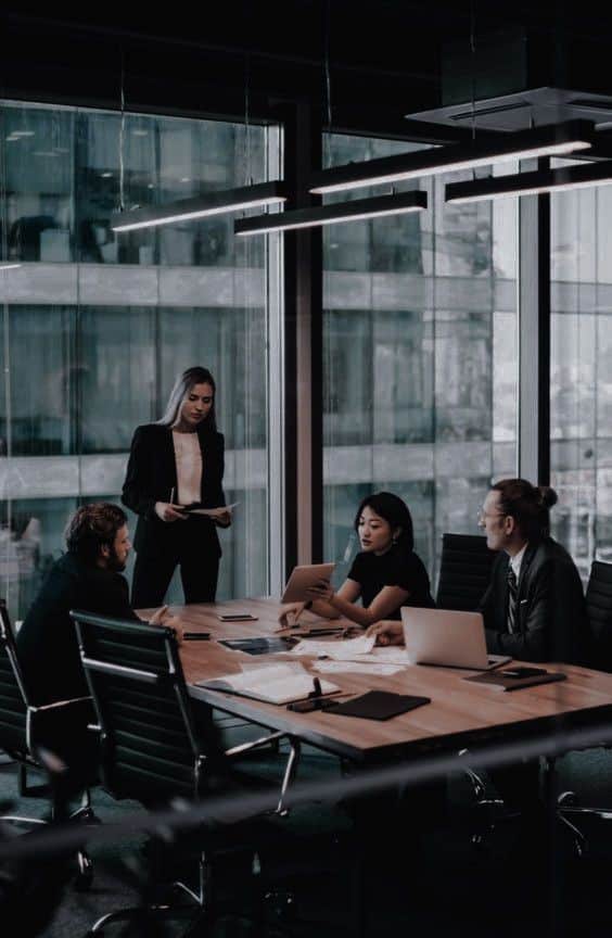 Business meeting in modern office with diverse team discussing strategy around a table, laptops and documents visible.