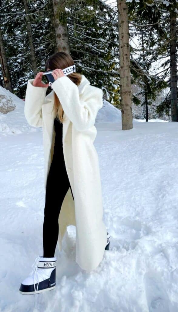 Fashionable woman in snowy forest, white coat and boots, covering face with branded visor. Winter style and luxury.