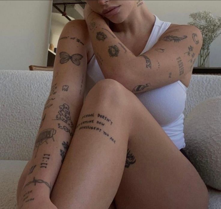 _ 14 Person sitting with multiple tattoos on arms and legs, wearing a white tank top, relaxing on a couch in a softly lit room. | Sky Rye Design Person sitting with multiple tattoos on arms and legs, wearing a white tank top, relaxing on a couch in a softly lit room.