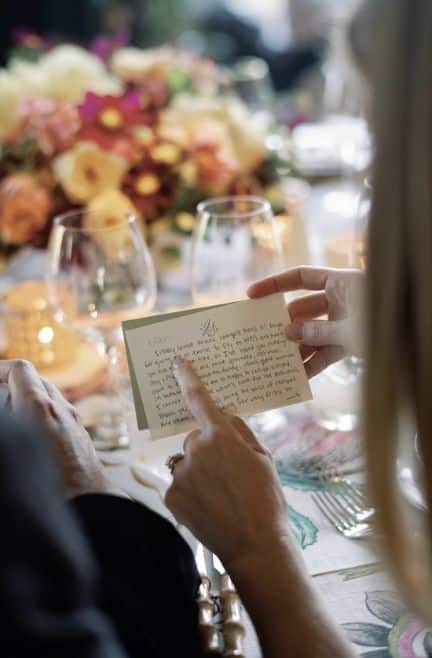 Screenshot Hands holding a handwritten card at an elegantly set table with flowers and wine glasses, creating a warm, festive atmosphere. | Sky Rye Design Hands holding a handwritten card at an elegantly set table with flowers and wine glasses, creating a warm, festive atmosphere.