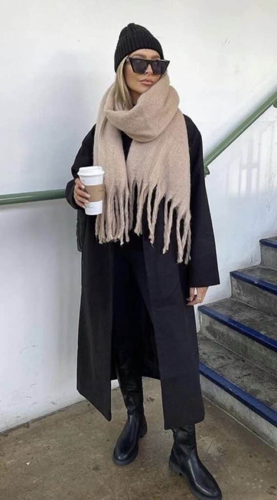 Woman in black coat, beige scarf, and beanie holding coffee cup. Winter fashion outfit on stairway.