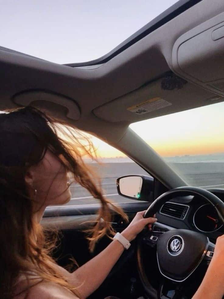 Person driving a Volkswagen, enjoying a sunset viewed through the car's sunroof, with hair blowing in the wind.