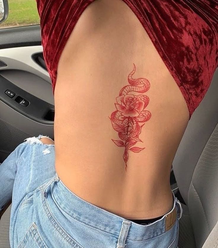 Tattoo of a rose with a snake on ribcage, person in red top and jeans sitting in a car, showcasing intricate body art.