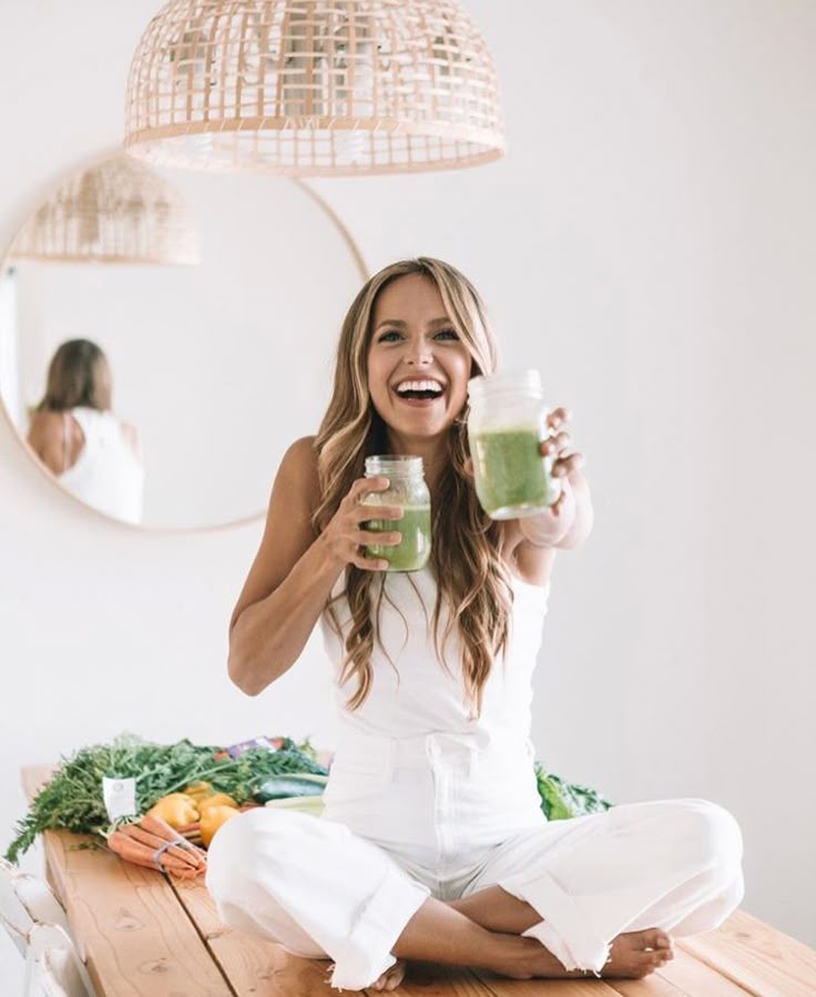 _ 15 Woman smiling and holding two green smoothies, sitting on table with fresh vegetables, under stylish pendant light. | Sky Rye Design Woman smiling and holding two green smoothies, sitting on table with fresh vegetables, under stylish pendant light.