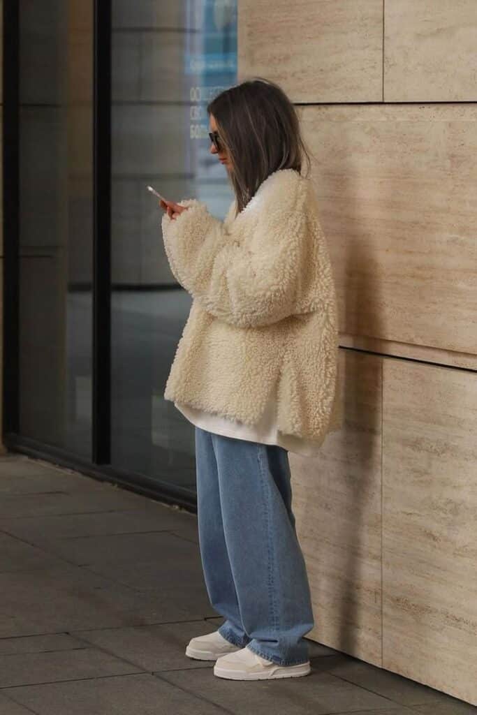 Woman in a cozy shearling jacket and jeans checks phone outdoors, urban style fashion.
