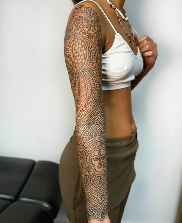 Woman with intricate full-arm mandala tattoo wearing a white top and brown pants.