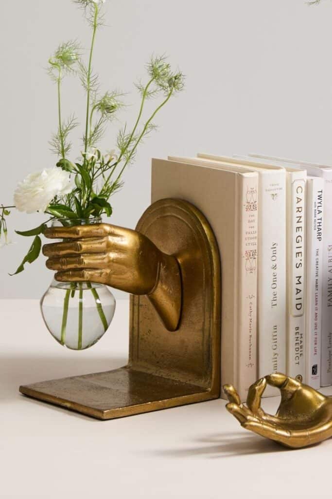_ 16 Elegant bookend with a golden hand holding a small vase with flowers, positioned next to a row of books for a stylish display. | Sky Rye Design Elegant bookend with a golden hand holding a small vase with flowers, positioned next to a row of books for a stylish display.