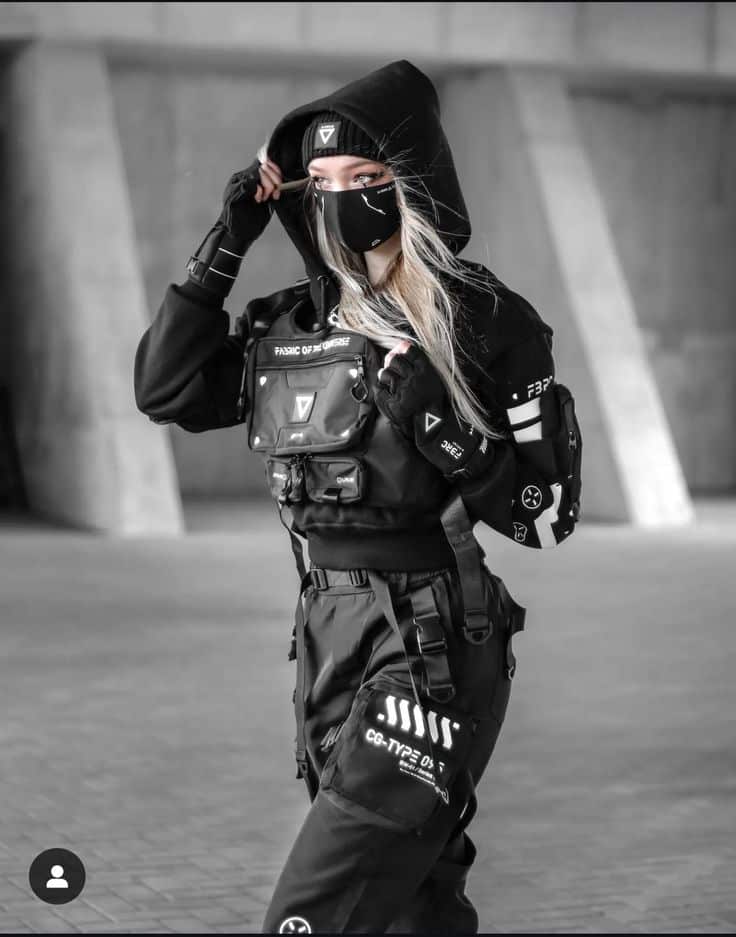 Person in black tactical gear and mask, with futuristic accessories, poses confidently in an urban setting, embracing a cyberpunk style.