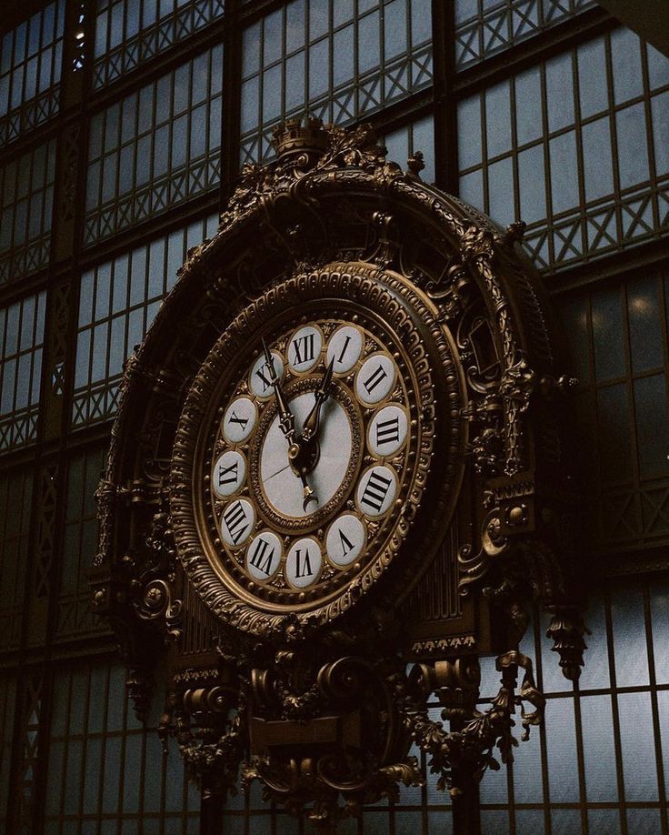 Ornate antique clock with Roman numerals, featuring intricate gold details, set against a large grid-windowed wall.
