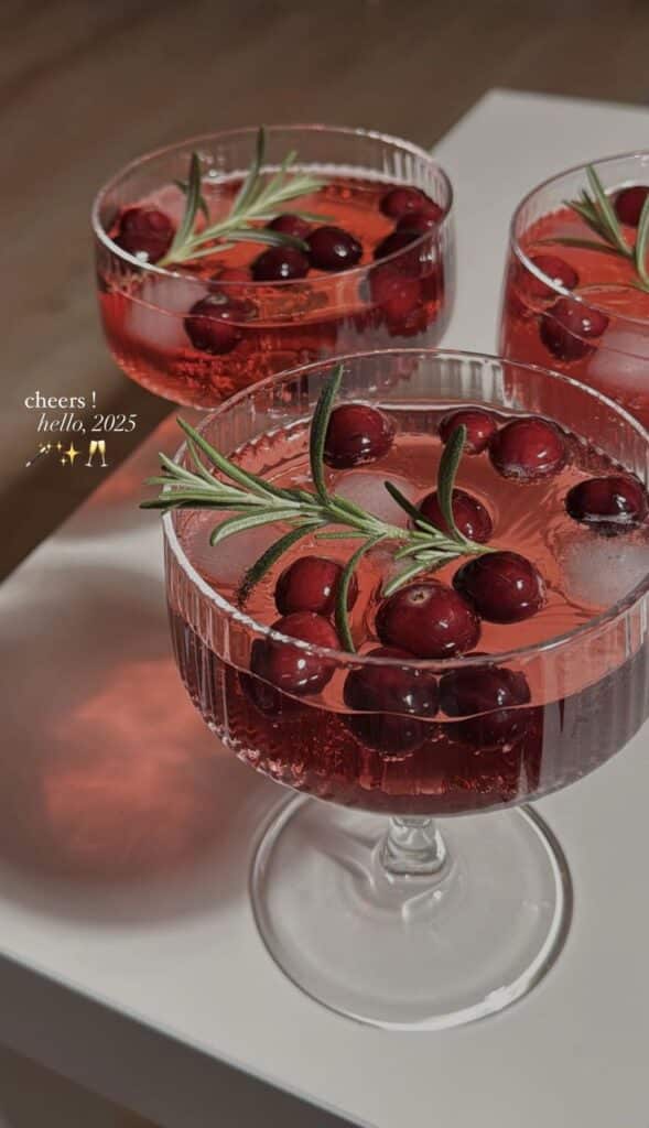 Festive cranberry cocktails with rosemary garnish, celebrating New Year 2025. Cheers to a sparkling start on a white table.
