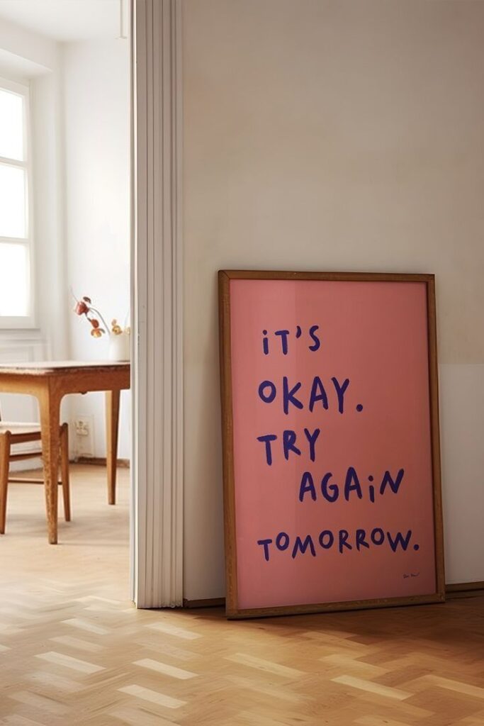 Framed quote art in a room: It's okay. Try again tomorrow. Pink background with blue text, leaning against a wall.