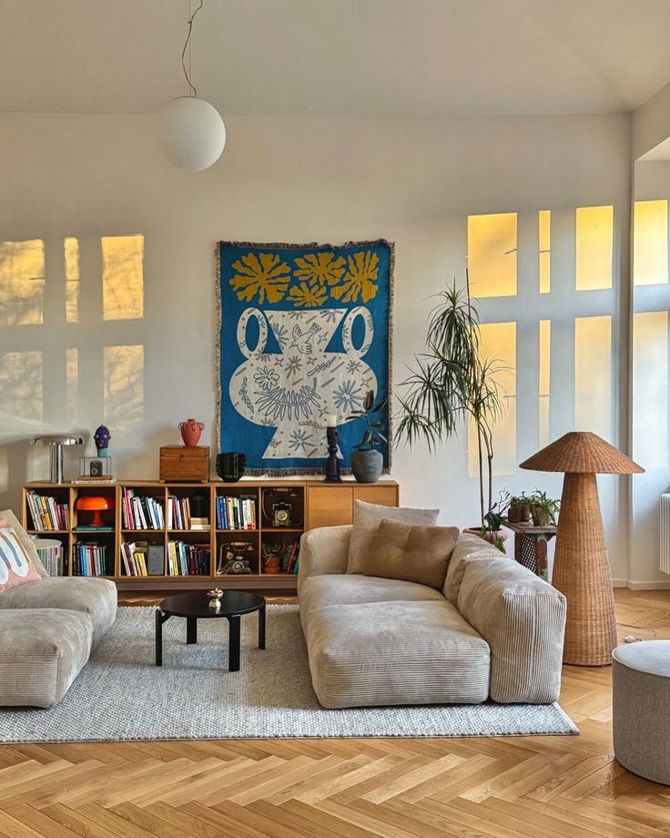 _ 16 Modern living room with cozy beige sofas, a vibrant blue wall art, wooden flooring, and bookshelf under warm lighting. | Sky Rye Design Modern living room with cozy beige sofas, a vibrant blue wall art, wooden flooring, and bookshelf under warm lighting.