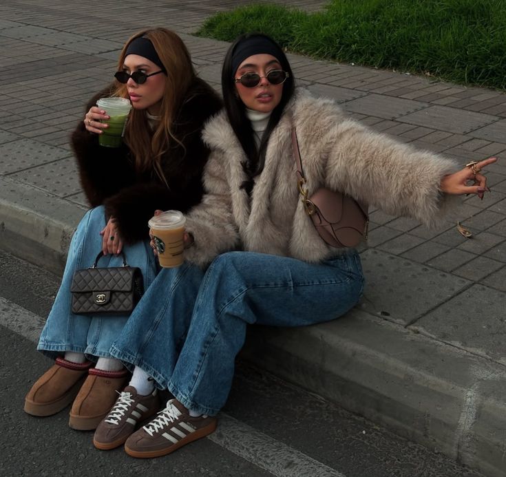 Two fashionable women in fur coats, jeans, and sunglasses sitting with drinks on a city sidewalk.