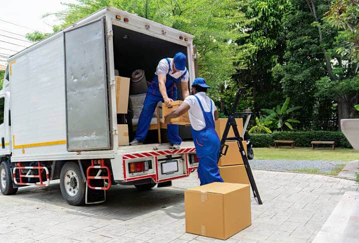 Movers in blue uniforms unloading cardboard boxes from a white truck in a leafy residential area.