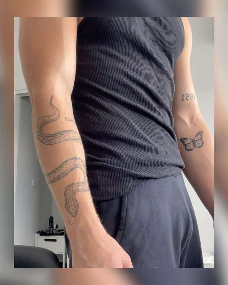 Man in black tank top showing tattoos of a snake and butterfly on arms in a casual setting.