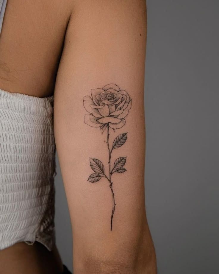 Minimalist black ink rose tattoo on upper arm, showcasing artistic floral design against a gray background. Perfect for tattoo inspiration.