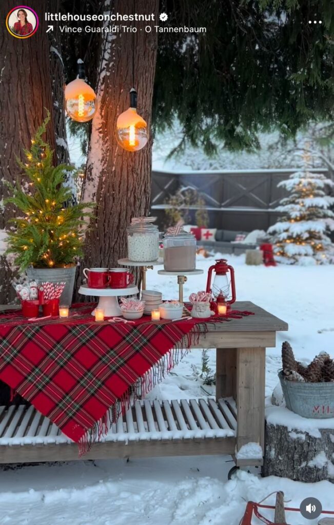 Cozy winter outdoor setup with festive plaid tablecloth, lanterns, hot cocoa, and candy canes, set against a snowy backdrop with lights.