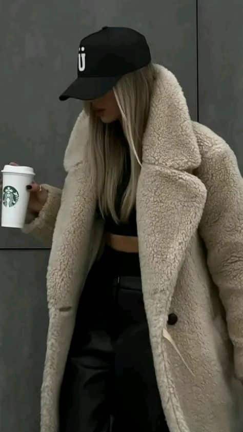 Stylish woman in a faux fur coat holding a Starbucks cup, wearing a black cap, epitomizing winter fashion elegance.