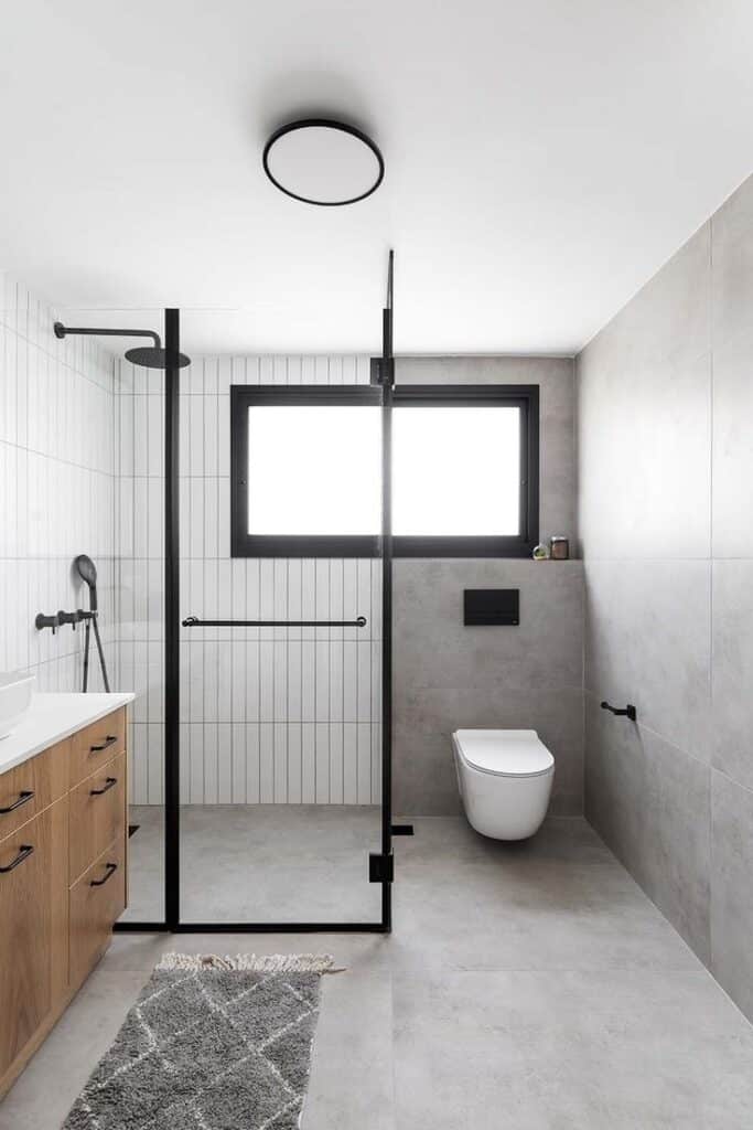 Modern bathroom with glass shower, wall-mounted toilet, wooden vanity, and large window for natural light.