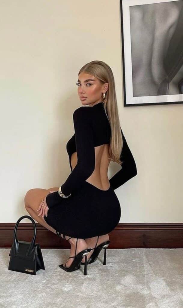 _ 18 Stylish woman in a sleek black backless dress and heels poses indoors with a black handbag and framed art on the wall. | Sky Rye Design Stylish woman in a sleek black backless dress and heels poses indoors with a black handbag and framed art on the wall.