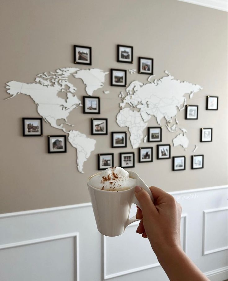 Hand holding a cappuccino in front of a world map photo wall, blending travel inspiration with cozy moments at home.