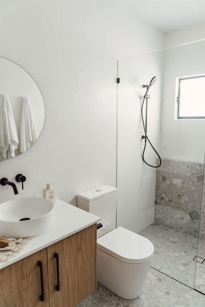 Modern minimalist bathroom with wood vanity, round mirror, white fixtures, and glass shower.