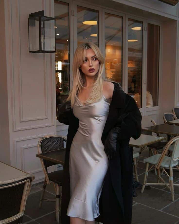 _ 19 Stylish woman in a silver dress and black coat standing in a chic outdoor café setting. Fashionable blonde with captivating look. | Sky Rye Design Stylish woman in a silver dress and black coat standing in a chic outdoor café setting. Fashionable blonde with captivating look.