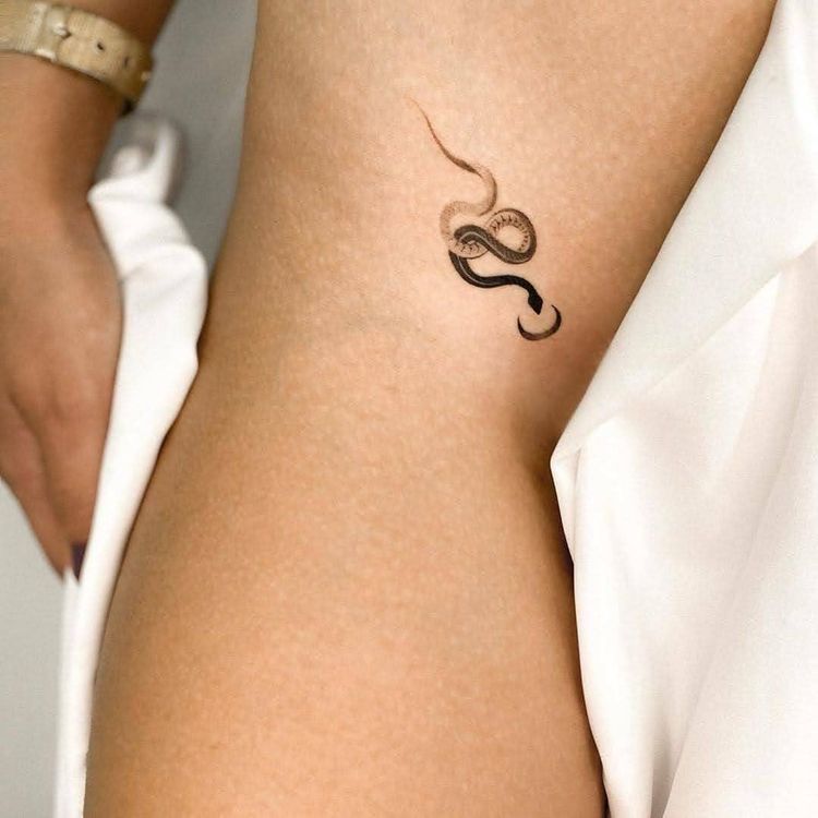 Elegant minimal snake tattoo on a person's thigh, highlighted by the softness of light skin tones and a white garment.