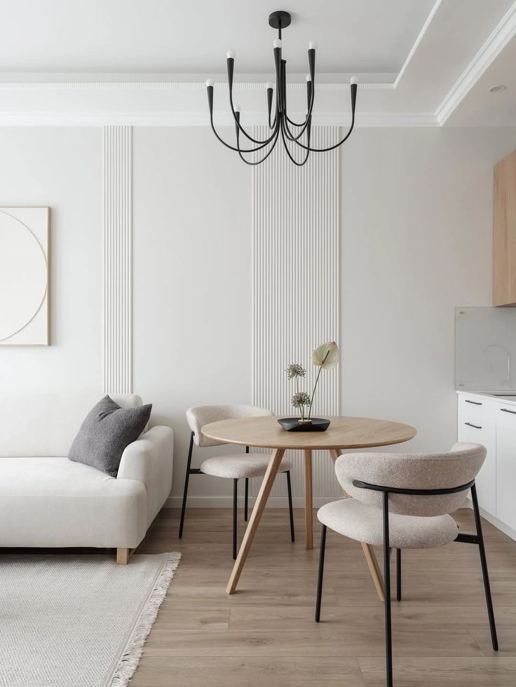 Minimalist living room with a round wooden table, beige chairs, white sofa, and modern black chandelier, enhanced by soft natural lighting.